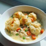 A close-up of a white bowl filled with creamy slow cooker chicken and dumplings, topped with golden biscuits and vegetables.