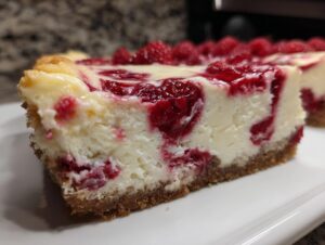 Close-up of a thick slice of creamy white chocolate cheesecake swirled with raspberry filling and topped with fresh raspberries.