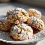 A stack of light yellow, crinkle-top cool whip cookies heavily dusted with powdered sugar on a white plate.