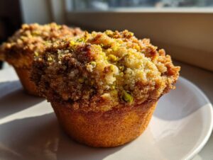 A close-up of a golden baked pistachio muffin with a sugary, crumbly topping and visible green pistachio pieces.