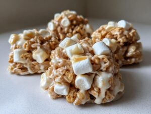 Four homemade avalanche cookies made with cereal, white chocolate, and mini marshmallows, glistening on a white surface.