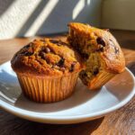 Two fluffy banana chocolate chip muffins, one whole and one broken open to show the texture, sitting on a white plate.