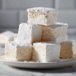 A stack of thick, square homemade marshmallows generously dusted with white powdered sugar on a white plate.