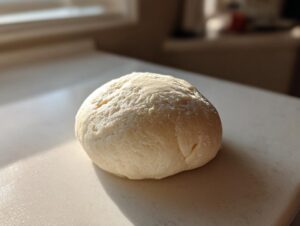 A smooth, light-colored ball of fresh marzipan recipe dough resting on a white surface in bright sunlight.