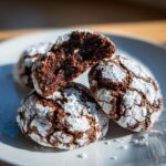 Close-up of rich, dark chocolate crinkle cookies dusted heavily with powdered sugar, one cookie broken open to show the fudgy interior.