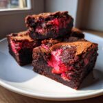 Close-up of fudgy strawberry brownies stacked on a white plate, showing bright pink strawberry filling inside the dark chocolate.