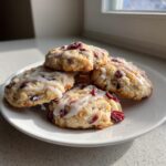 A stack of four freshly baked cranberry orange cookies drizzled with white glaze, sitting on a white plate near a window.