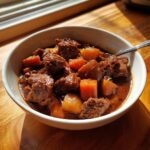 Close-up of a white bowl filled with rich, dark 8 hour pork stew chunks and carrots, sitting on a wooden table.