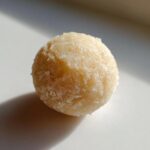 A single, round, pale yellow marzipan ball dusted with fine sugar, sitting on a white surface.