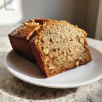 A thick slice of moist banana bread recipe showing walnuts embedded in the crumb, served on a white plate.