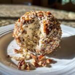 A creamy cheese ball recipe covered in chopped pecans, with a scoop removed to show the texture inside, resting on a white plate.