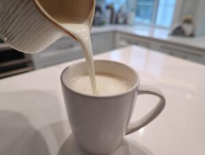 Milk being poured from a ceramic pitcher into a white mug to make white hot chocolate.