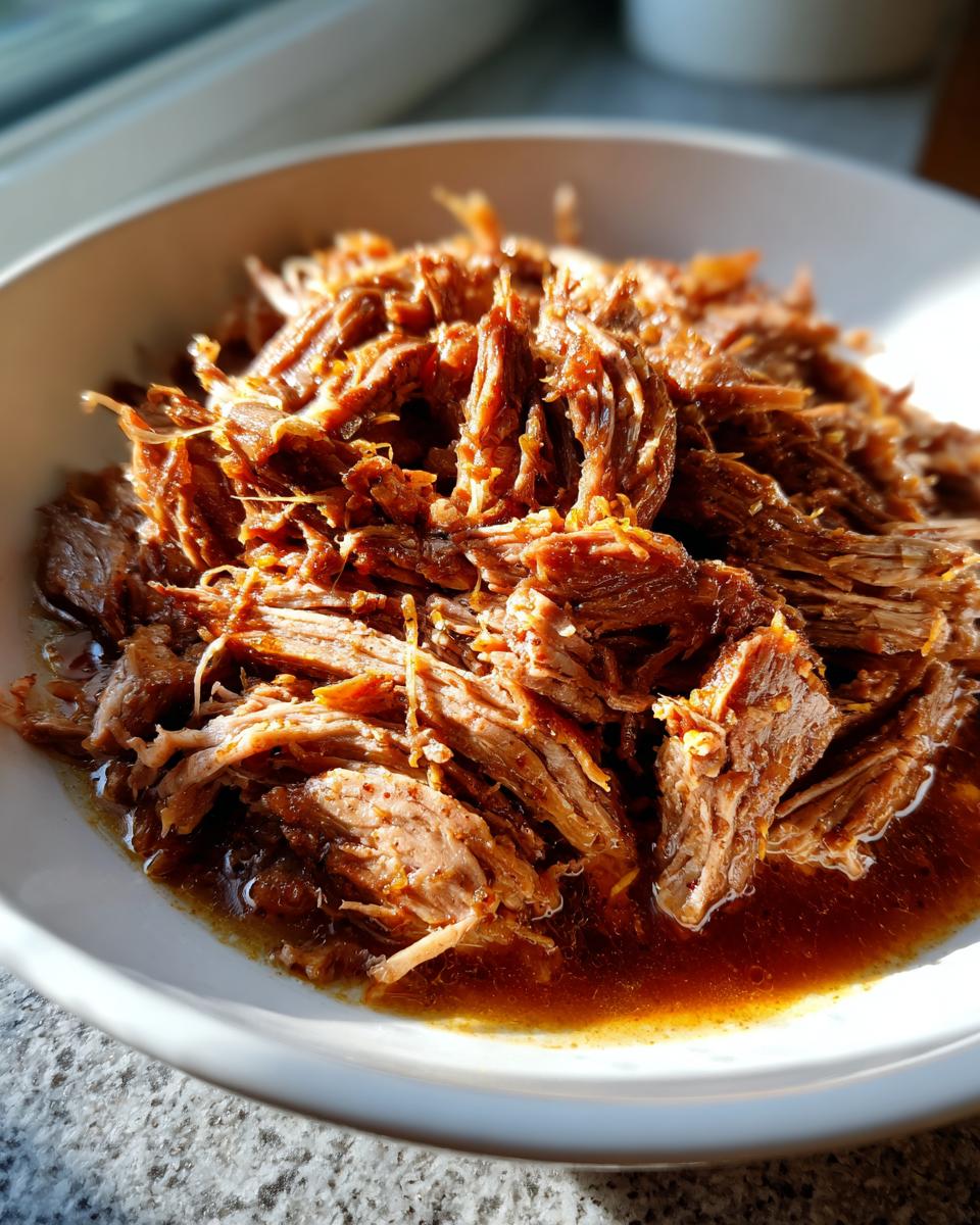 Close-up of tender, shredded Mississippi pot roast sitting in its rich brown juices in a white bowl.