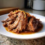 Close-up of tender, shredded Mississippi Pot Roast served on a white plate with rich gravy and a garnish of zest.