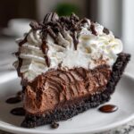 A decadent slice of chocolate ice cream pie featuring a dark crust, rich chocolate filling, and topped with whipped cream and cookie crumbles.