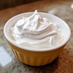 A close-up of thick, glossy white royal cookie icing piped into soft peaks in a small yellow bowl.