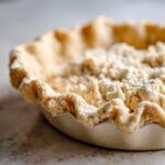 Close-up of an unbaked, fluted butter pie crust dough in a white pie dish, showing crumbly texture.
