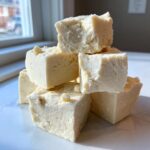 A stack of creamy, pale squares of homemade white chocolate fudge garnished with small white chocolate chips.
