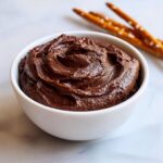 A close-up of a thick, rich chocolate dip swirled in a white bowl, with pretzel rods visible in the background.