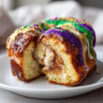 A cross-section view of a rich, cinnamon-swirled king cake topped with purple, green, and gold icing and sprinkles.