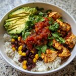 A close-up view of a vibrant chicken burrito bowl featuring seasoned chicken, white rice, black beans, corn, salsa, avocado slices, and cilantro.