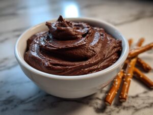 A close-up of a creamy, dark chocolate dip swirled high in a white bowl, served next to salty pretzel sticks.