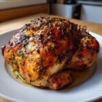 A whole, perfectly glazed and seasoned roasted chicken resting on a white plate with pan juices.