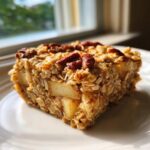 A close-up of a hearty slice of baked oatmeal, featuring visible apple chunks and topped with pecans.