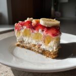 A delicious slice of banana split cake showing layers of graham cracker crust, cream filling, pineapple, strawberries, and banana slices.