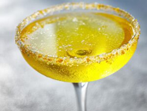Close-up of a bright yellow lemon drop cocktail in a coupe glass with a coarse sugar and zest rim.