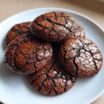 A stack of five rich, dark chocolate brownie cookies with distinctively cracked tops on a white plate.