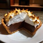 A slice removed from a small chocolate meringue pie, showing the rich dark filling and tall, torched meringue topping.