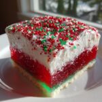 A square slice of Christmas poke cake showing layers of cake, red gelatin, white topping, and red/green sprinkles.