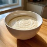 A creamy, smooth bowl of cauliflower soup garnished with spices, sitting on a wooden surface in natural light.