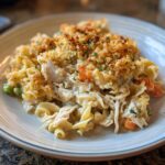 A serving of creamy chicken noodle casserole featuring egg noodles, shredded chicken, peas, carrots, and a golden breadcrumb topping.