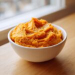 A close-up of creamy, bright orange mashed sweet potatoes served in a white bowl.