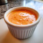 Close-up of a creamy, orange-colored burger sauce speckled with red spices, served in a white fluted ramekin.