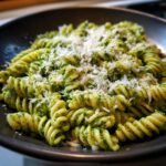 A close-up of fusilli pasta coated in bright green sauce, served as creamy pesto pasta and topped with grated Parmesan.