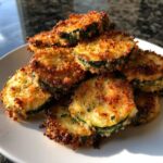 A pile of golden brown, crispy baked parmesan zucchini slices stacked on a white plate.