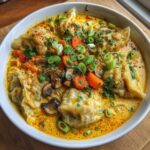 A close-up of dumplings served in a rich, yellow coconut curry soup, topped with mushrooms, carrots, and green onions.