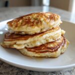 A close-up of a stack of three fluffy banana pancakes drizzled with syrup on a white plate.