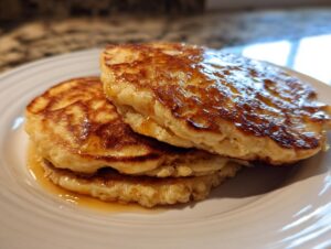 A close-up of three fluffy banana pancakes stacked on a white plate, drizzled generously with syrup.