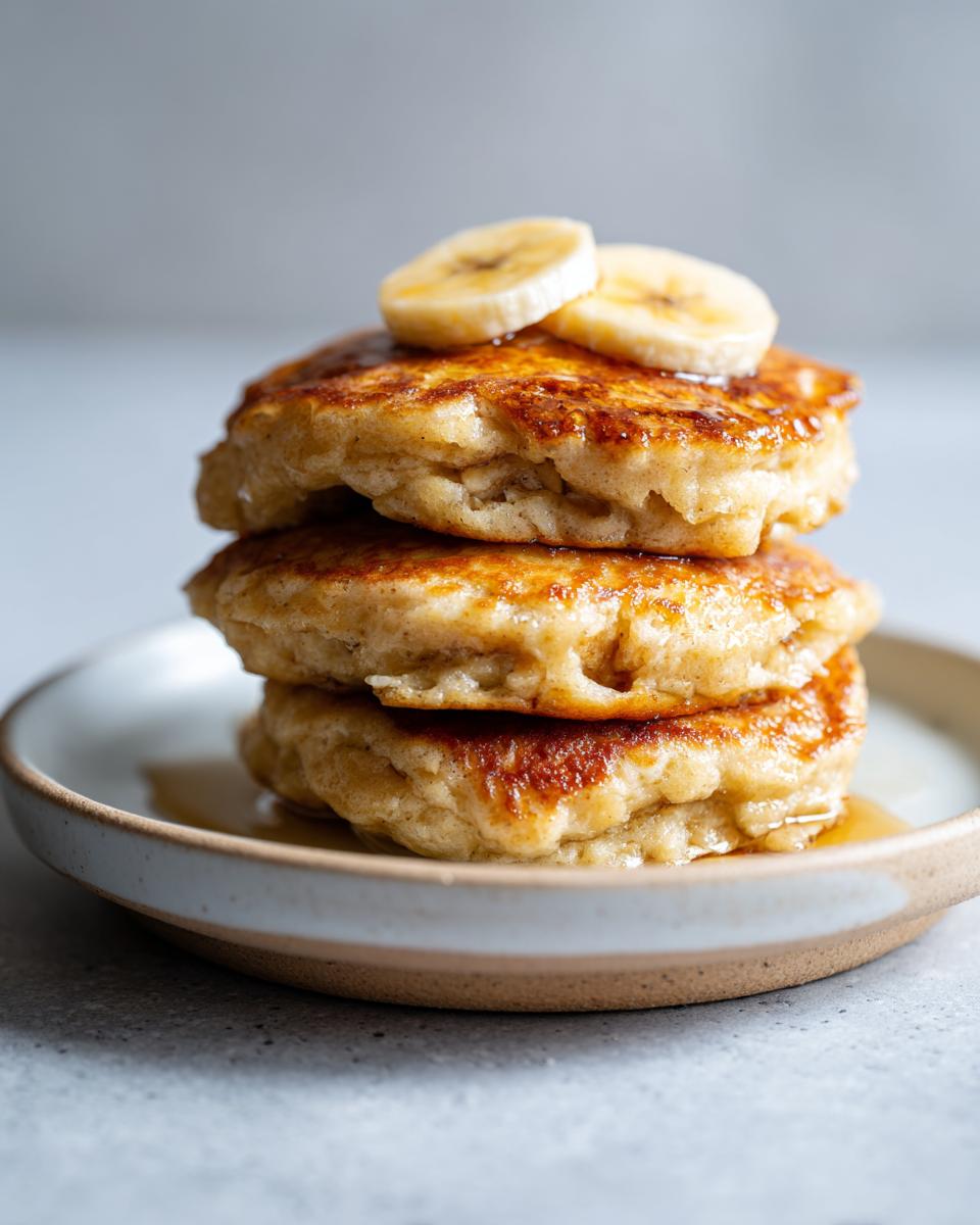 A close-up of a stack of three fluffy banana pancakes drizzled with syrup and topped with fresh banana slices.