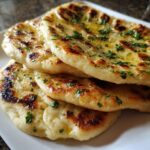 Close-up of a stack of three fluffy naan bread recipe pieces, brushed with melted butter and sprinkled with fresh parsley.