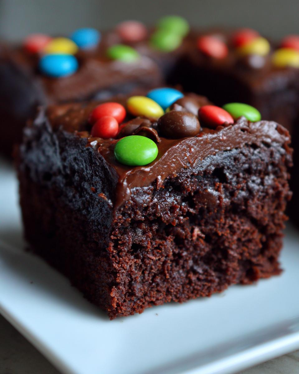 A close-up of a rich, fudgy square of cosmic brownies topped with chocolate frosting and colorful candy pieces.