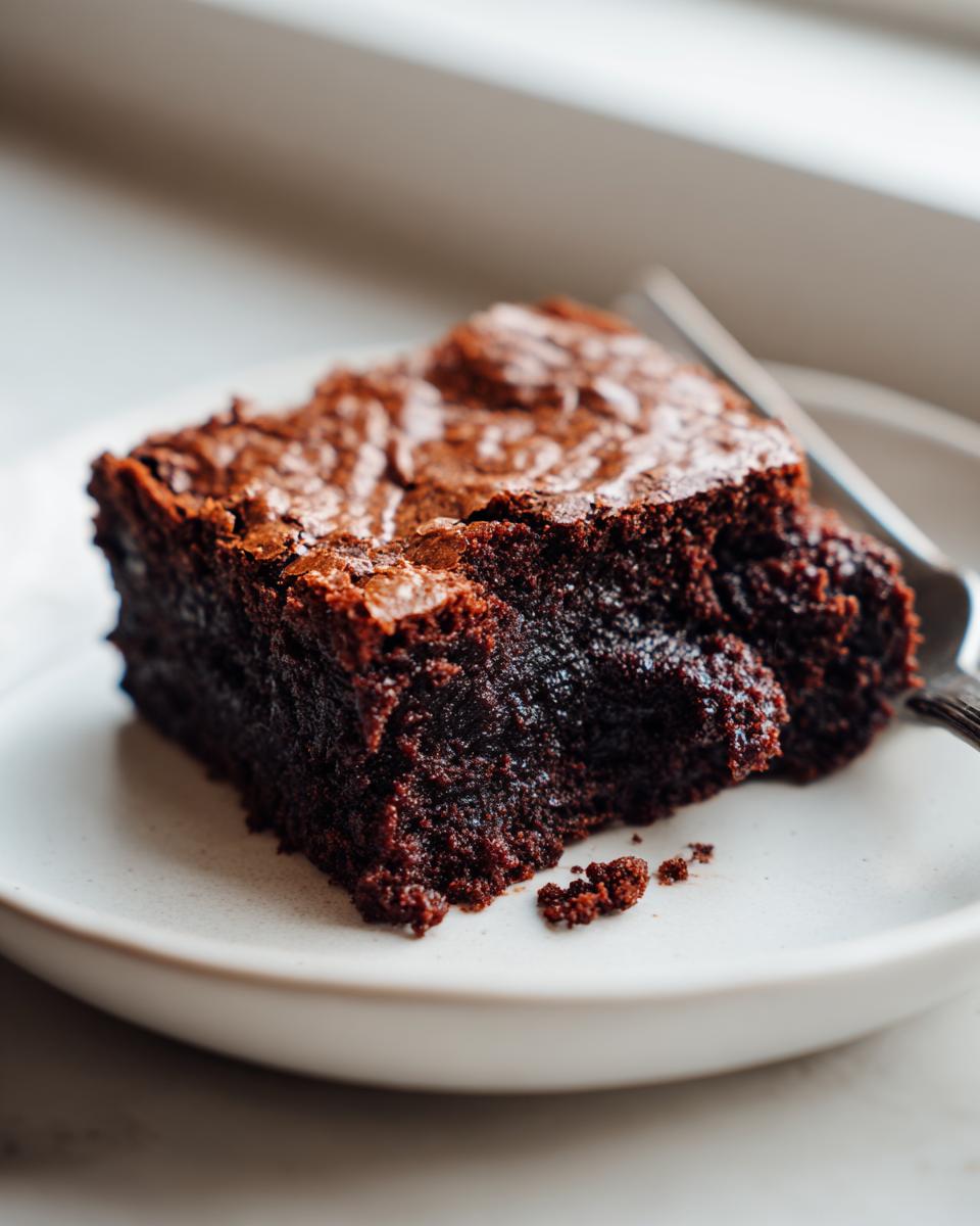 Close-up of a fudgy, dark chocolate slice of gingerbread brownies on a white plate with a fork.