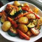 A close-up of a white bowl filled with golden brown Garlic Herb Roasted Veggies including baby potatoes, carrots, and zucchini.