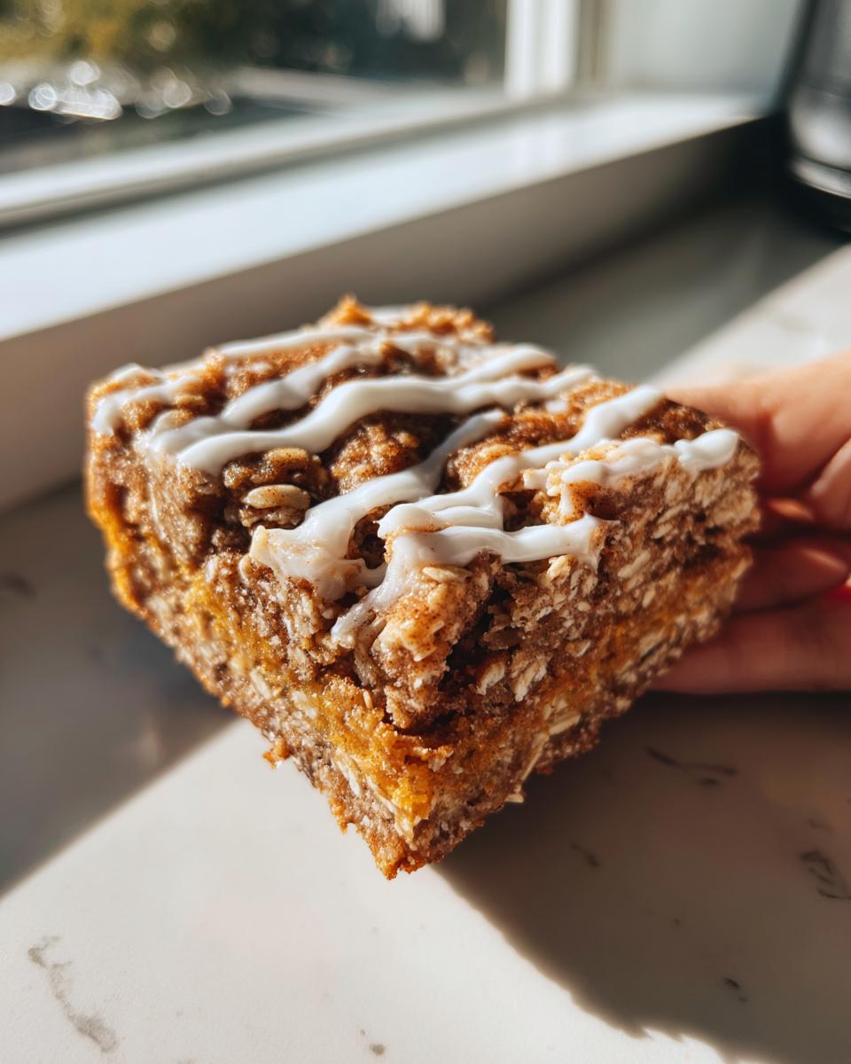 A hand holding a thick, square oatmeal bars slice topped with white icing drizzle, backlit by a window.