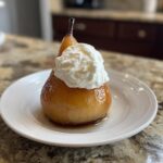A perfectly glazed poached pear topped with a dollop of whipped cream, served on a white plate.