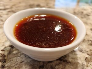 Close-up of glossy, dark brown homemade teriyaki sauce in a small white bowl on a granite countertop.